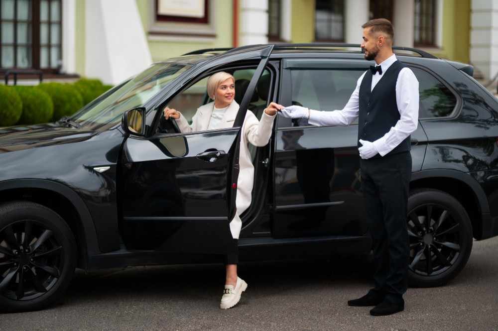 A professional chauffeur helps a woman step out of a black luxury car outside a building, offering courteous assistance during a pickup.