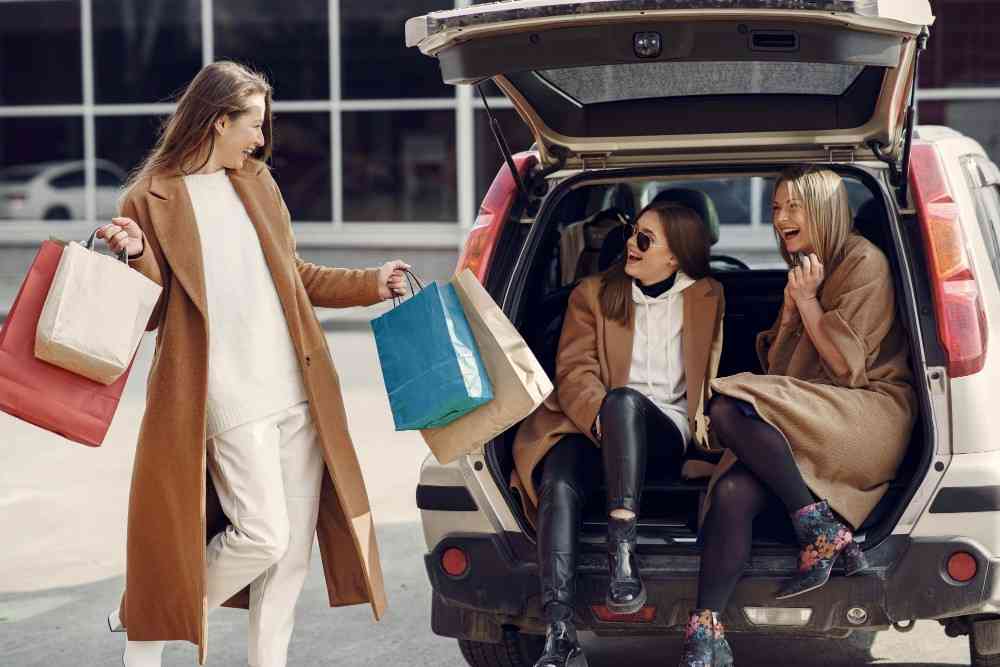 Friends returning from a shopping trip with bags, sitting in the back of a car and smiling together.